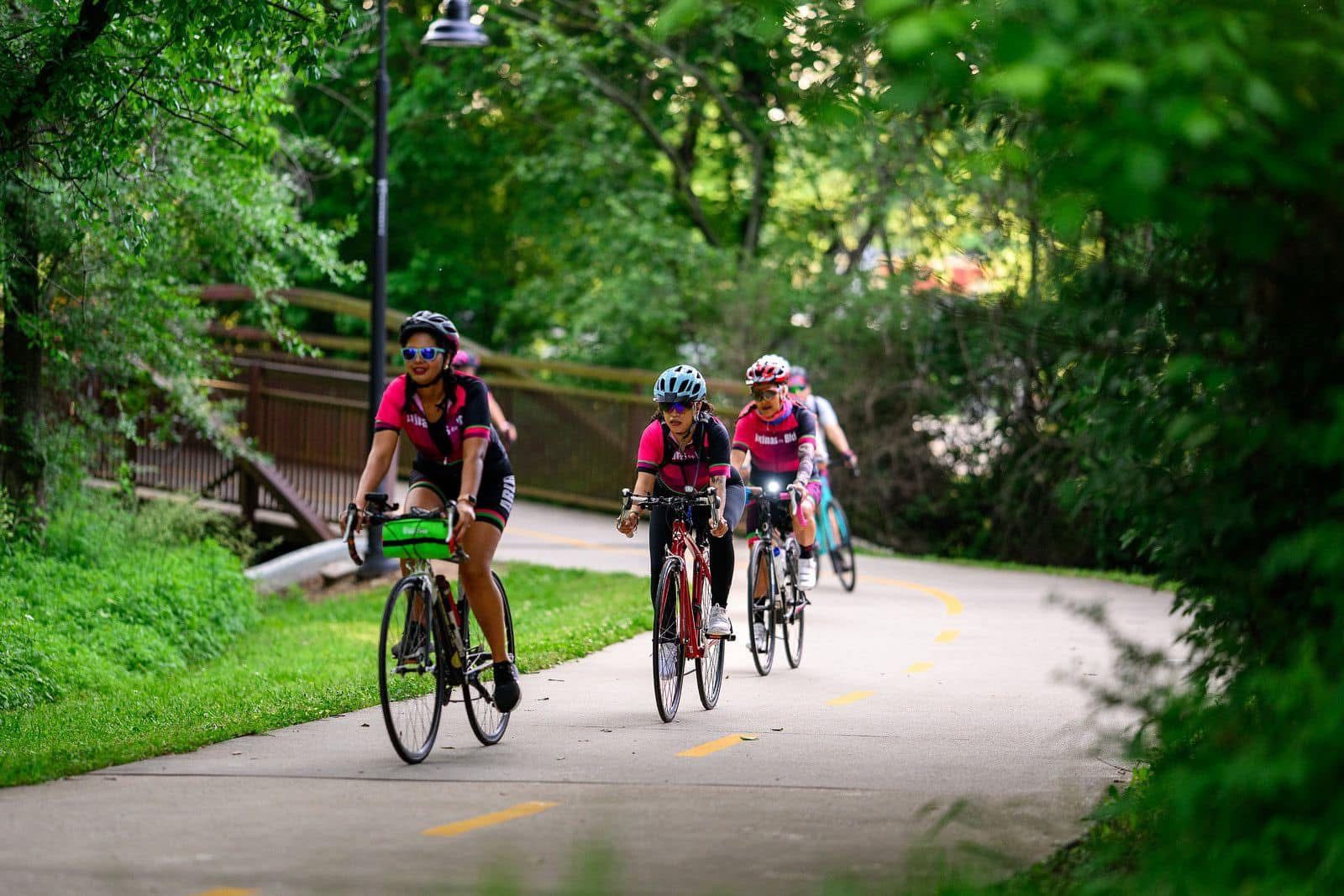 Latinas en Bici members in matching magenta jerseys riding together on a beautiful tree-lined greenway trail in Northwest Arkansas